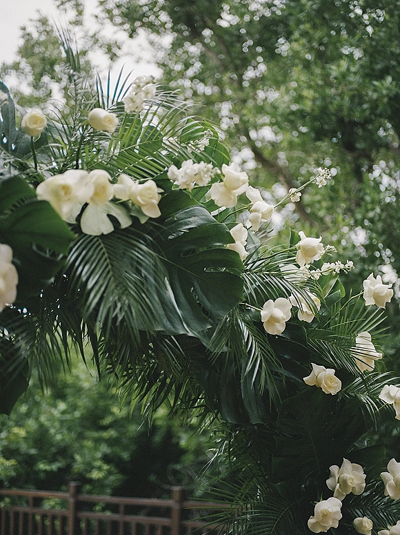 Wedding floral arch with tropical wedding arch styling, white roses and orchids draped over monstera leaves on a garden railing backdrop