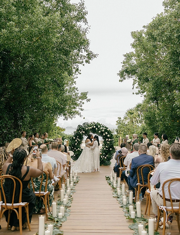 Ceremony kiss between two brides under a round white-flower arch with greenery garland and candle-lined aisle on an oceanfront deck