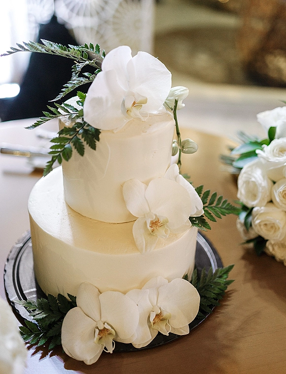 Wedding cake with white orchids and fern greenery on a cake stand, set on a wooden table beside place settings and florals