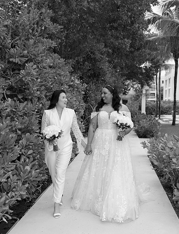 Couple portrait of two brides holding hands and walking on a sidewalk, one in lace dress and one in jumpsuit, holding a rose bouquet