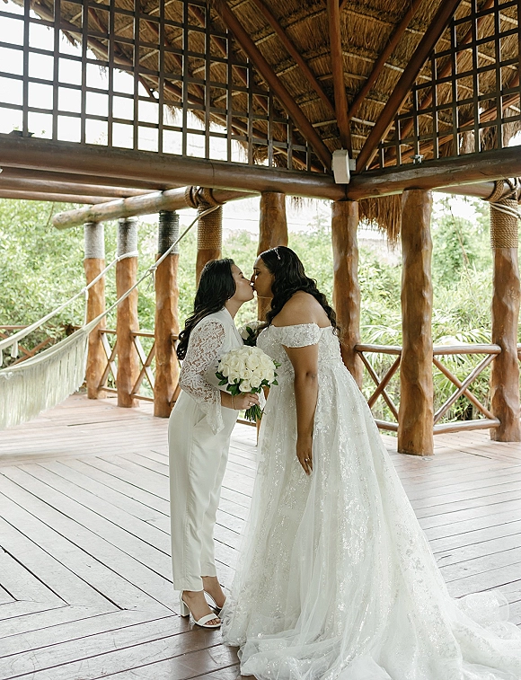 Wedding kiss portrait of two brides sharing a kiss, one holding a white rose bouquet under a thatched wooden pavilion with tropical greenery