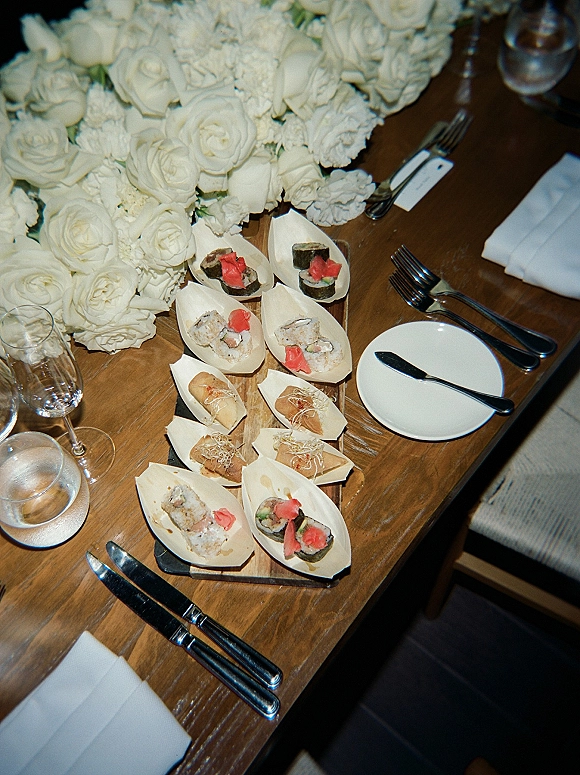 Wedding cocktail hour food with passed hors d'oeuvres wedding display of sushi rolls and finger sandwiches beside a white rose centerpiece on a wood table