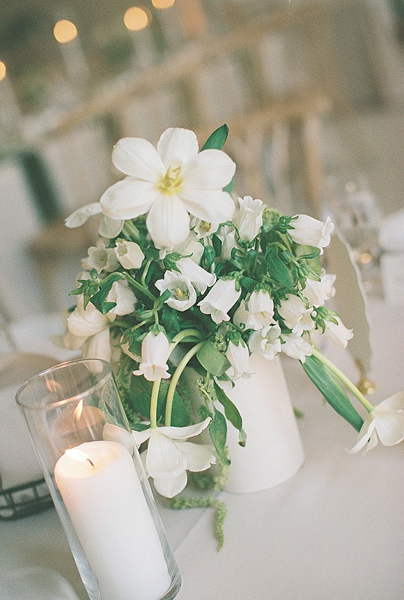 Wedding centerpiece with a white flower centerpiece in a bud vase beside a pillar candle in a glass hurricane on linen table under bistro lights