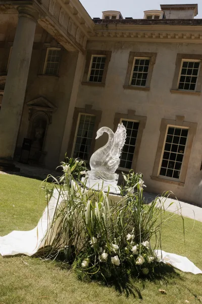 Wedding decor installation featuring an ice swan sculpture with white draping, greenery, and florals on a manor lawn backdrop
