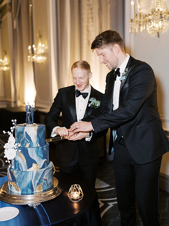 Cake cutting moment as two grooms in tuxedos hold a knife beside a three-tier cake with topper figurines in a chandelier-lit ballroom