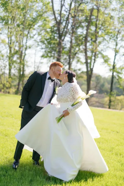 Wedding kiss portrait of groom dipping the bride for an outdoor wedding kiss, her lace dress and veil flowing on a sunny lawn with trees