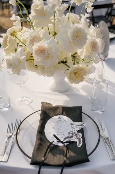 Reception tablescape with white rose centerpiece in a pedestal vase, black napkin place setting, menu and place cards, sunlit outdoor seating behind