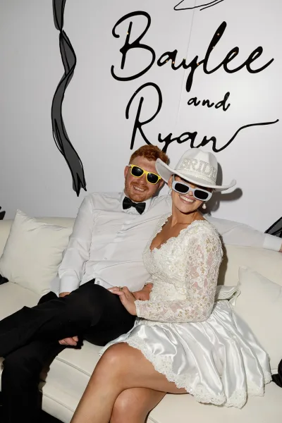 Couple portrait in a wedding photo booth, bride in cowboy hat and sunglasses beside groom on a couch against a white wall backdrop