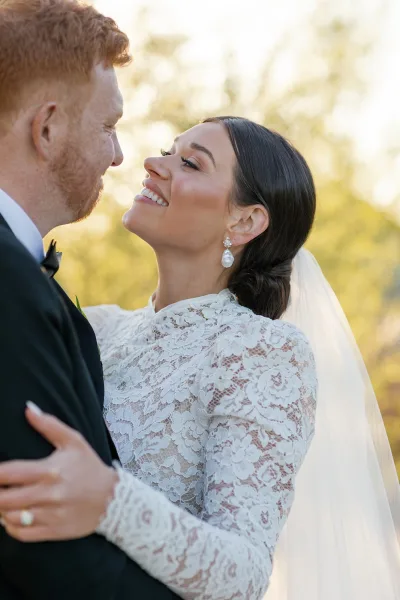 Couple portrait of bride and groom embrace, smiling forehead to forehead in golden hour light with cathedral veil against trees