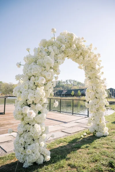 Wedding ceremony arch covered in white flowers, roses and hydrangeas, framing a lakeside dock with railing and blue sky beyond