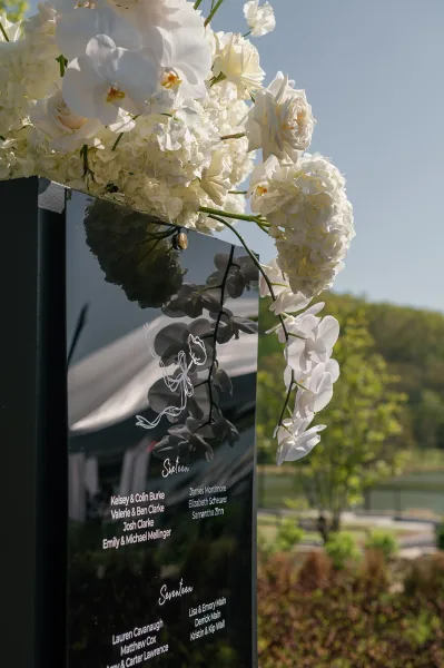 Wedding seating chart acrylic sign with white orchid and rose florals, framed by hanging blooms against trees and distant hills backdrop