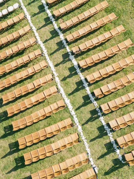 Ceremony seating with wood chairs arranged in rows around a double aisle lined with white flower petals on a grass lawn outdoors