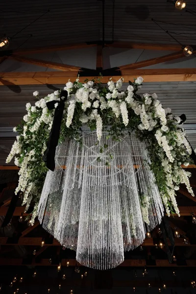 Wedding chandelier with white flowers and greenery, black ribbon accents, beaded fringe, and string lights under wood beams and a metal ceiling