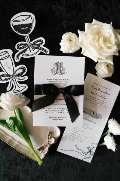 Invitation suite with black and white wedding invitations tied in a black satin bow, styled with white roses on a black fabric backdrop