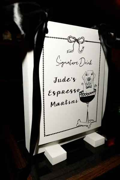 Signature drink sign on a small easel with a black ribbon bow, featuring a dog and martini illustration on a dark wood tabletop backdrop