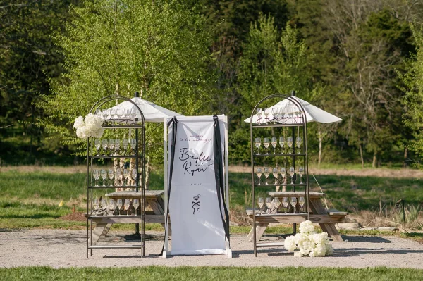 Champagne station with a wedding champagne wall of flutes on metal shelves, black ribbon and hydrangeas, set on a grassy park lawn under umbrellas