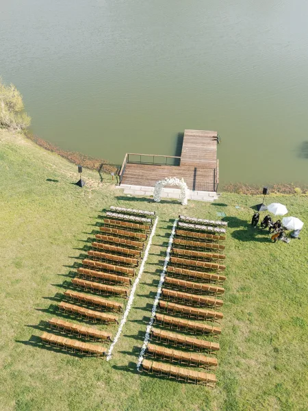 Ceremony setup with outdoor ceremony seating in wood chairs flanking a flower-lined aisle leading to a floral arch by the lake and dock
