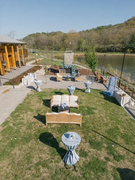Cocktail hour setup with wedding lounge seating, blue floral accents on cocktail tables under string lights by a lakeside patio fireplace
