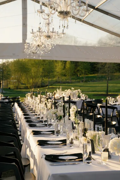 Reception tablescape with a long banquet table wedding setup, black taper candles and white florals under crystal chandeliers in a clear tent at sunset