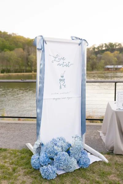 Wedding signage welcome sign banner with calligraphy lettering and a blue satin bow, framed on a stand beside hydrangeas by the lake railing