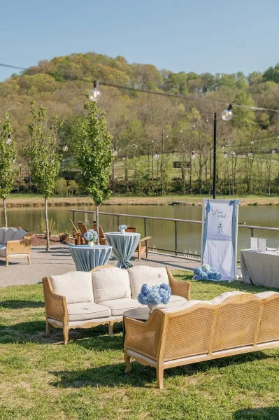 Outdoor lounge area with wedding lounge seating, sofas and cocktail tables with hydrangeas under string lights beside a lakeside lawn