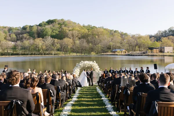 Ceremony moment at an outdoor wedding ceremony with bride and groom under a white floral arch on a lakeside lawn as guests watch