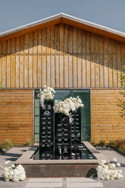 Wedding seating chart with black acrylic panels and orchid arrangements beside a water feature with floating candles outside a barn window