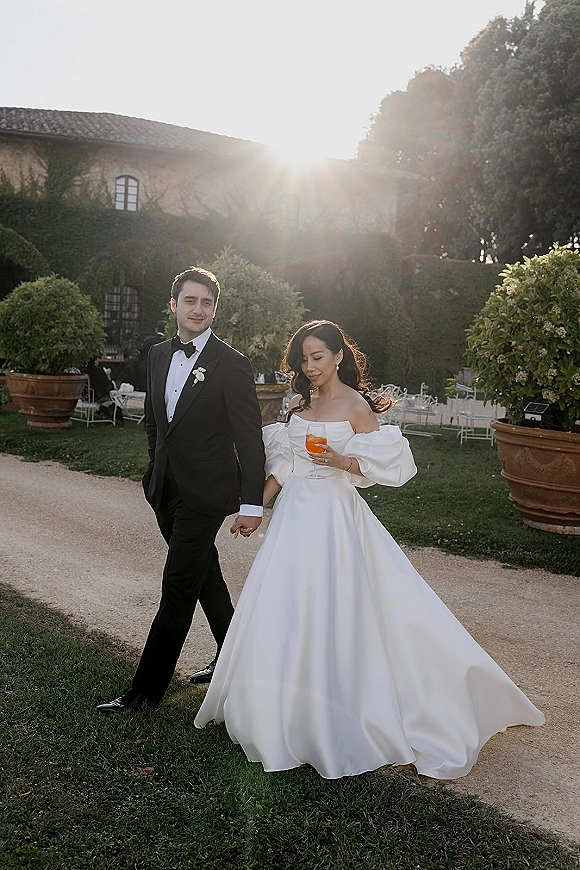 Couple portrait of bride and groom walking hand in hand, she holds a cocktail, on a sunlit garden path by a stone villa.