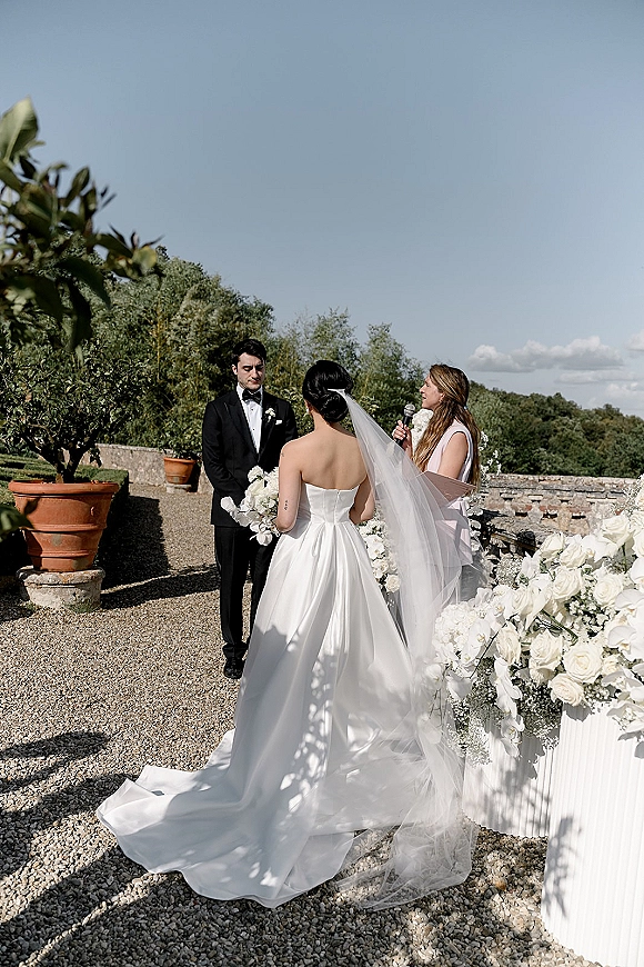 Wedding vows as bride in strapless satin dress and cathedral veil faces groom in black tux on a terrace with stone wall and sky backdrop