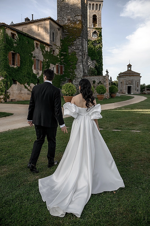 Couple portrait of bride and groom walking away hand in hand, her off-the-shoulder gown train flowing by an ivy-covered stone chapel path.