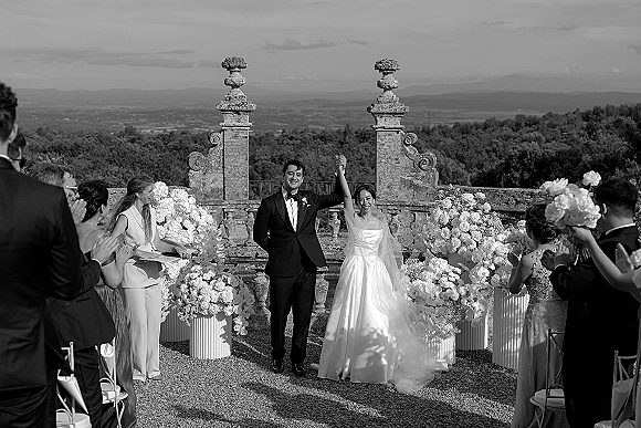 Recessional moment as bride and groom hold hands walking past ceremony chairs, bouquet and veil flowing on a stone terrace with mountain view