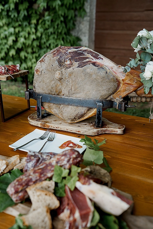 Wedding charcuterie table with prosciutto carving station, cured meats and bread on wood board, styled with white flowers and eucalyptus outdoors