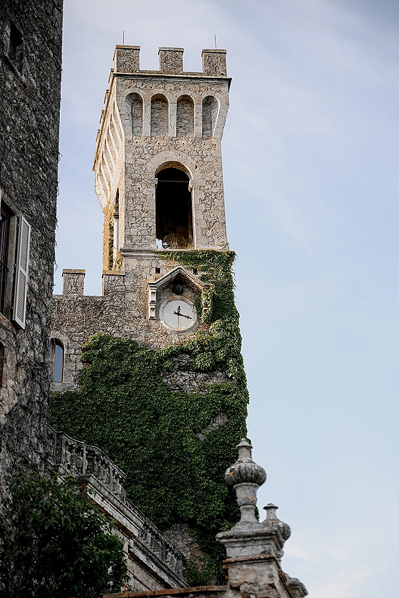 Castle exterior with ivy climbing a stone facade, featuring a clock tower and arched openings under a clear blue sky