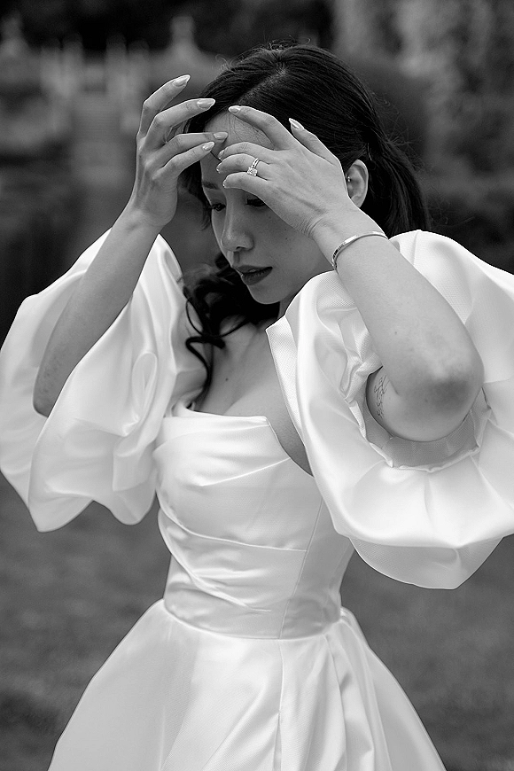 Bridal portrait in black and white of a bride adjusting her hair, showing an engagement ring and puff sleeve gown in a garden setting