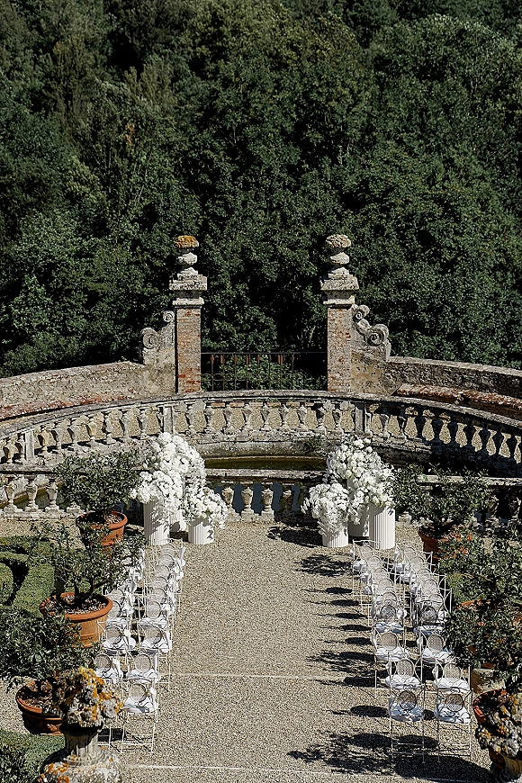 Ceremony setup for an outdoor wedding ceremony with white floral pedestals and cushioned aisle chairs on a stone terrace by trees