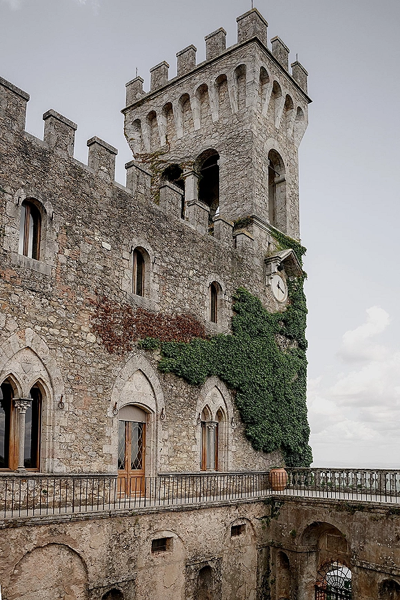 Castle wedding venue with ivy-covered stone tower and clock face, arched windows and wooden door above a stone courtyard under overcast sky