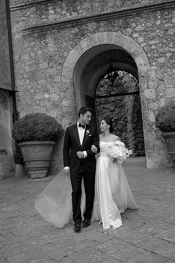 Couple portrait of bride and groom walking arm in arm, she holds a white orchid bouquet beneath a stone archway courtyard with ivy wall