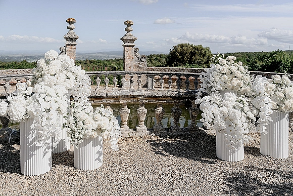 Ceremony floral arrangements of white roses, orchids and baby's breath in urns on ribbed pedestals along a stone terrace by a reflecting pool