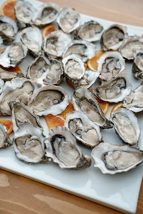 Oyster platter for a wedding raw bar with raw oysters on the half shell and orange slices on a white serving platter on a wood table