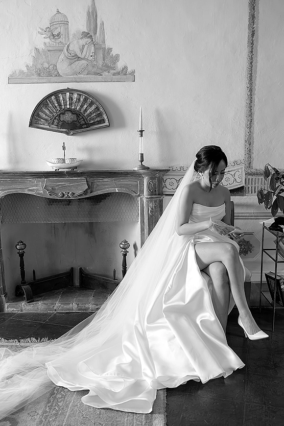 Bridal portrait in black and white of a bride sitting in a strapless gown and veil, reading a letter by a fireplace mantel with candles