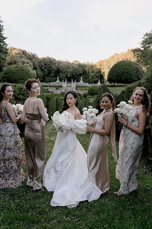 Bride with bridesmaids holding white rose bouquets, looking back over their shoulders on a stone staircase in a formal garden setting