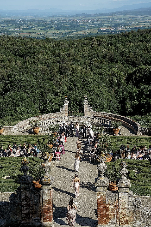 Outdoor wedding ceremony in a terraced garden with bridesmaid processional, white chairs and floral aisle runner, overlooking valley hills