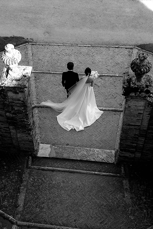 Couple portrait in an overhead wedding photo, bride in strapless gown with long veil holding bouquet beside groom in tux on stone staircase