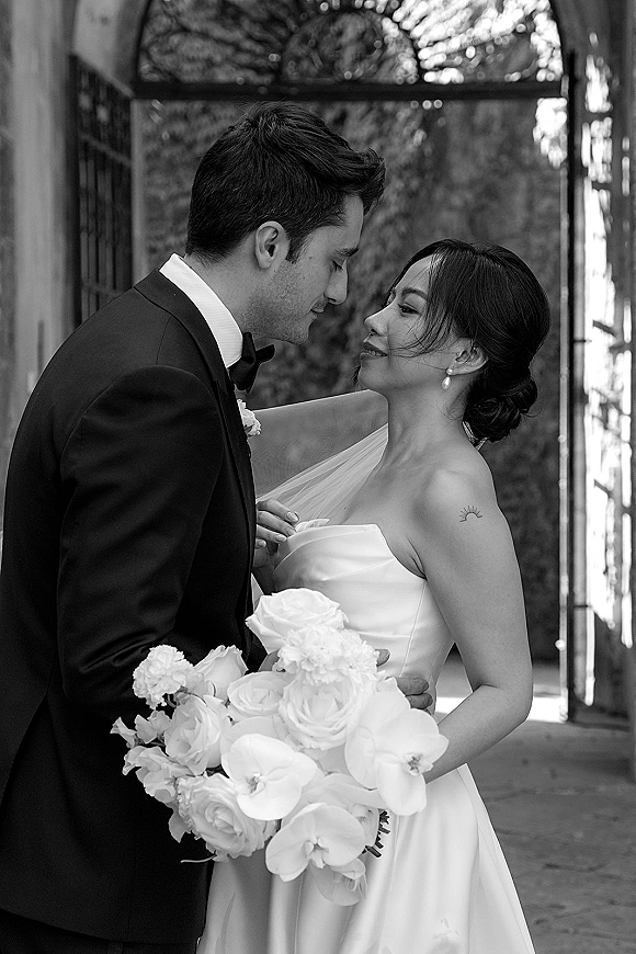 Wedding couple portrait in black and white, bride and groom touching foreheads as she holds an orchid bouquet by a wrought iron gate