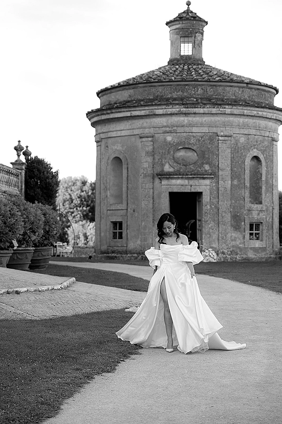 Bridal portrait of a bride in an off the shoulder wedding dress with puff sleeves and a flowing train on a gravel path by a stone pavilion