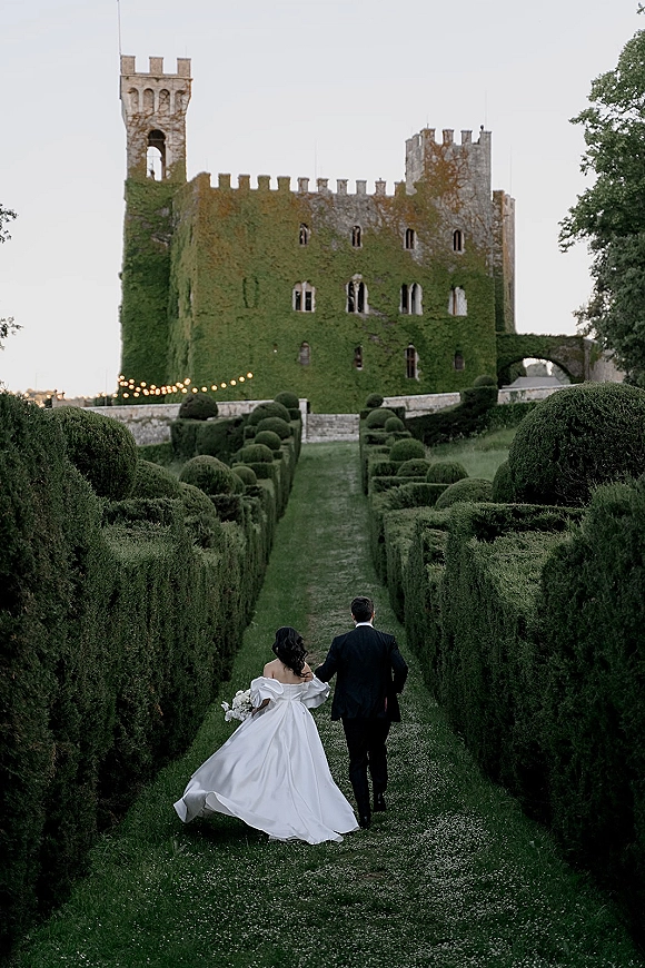 Couple portrait of newlyweds holding hands from behind, bride in ball gown with bouquet under string lights by ivy-covered castle walls