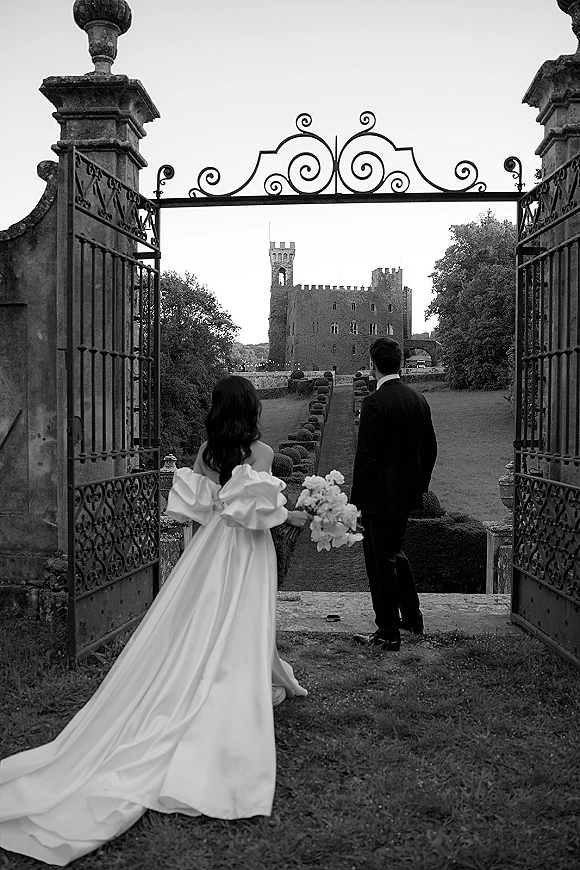 Couple portrait of bride and groom from behind, walking away through a wrought iron gate toward a castle garden path, bride holding bouquet