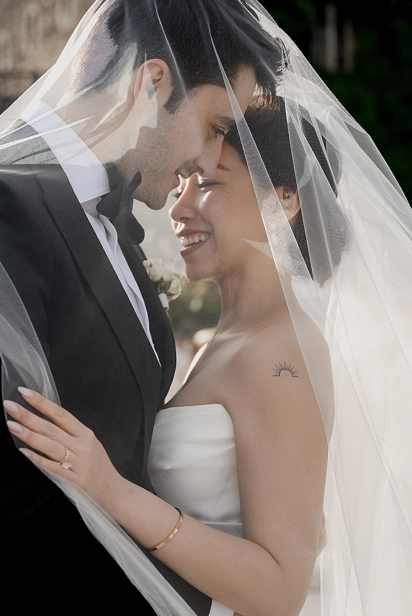 Wedding couple portrait of bride and groom under veil, touching foreheads as she smiles in a strapless dress against outdoor greenery