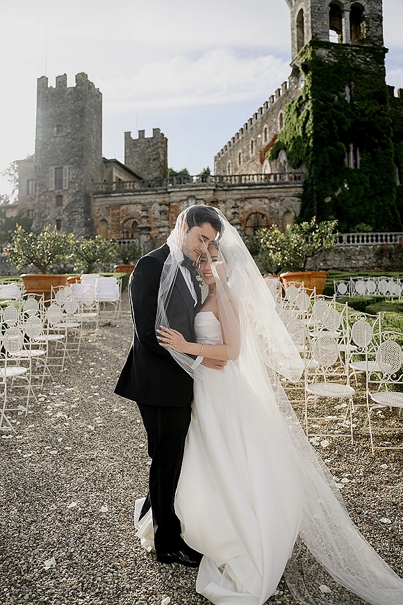Couple portrait of bride and groom embrace under veil, bride in strapless gown, with ivy-covered stone castle courtyard behind