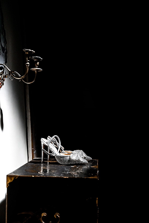 Wedding shoes, silver high heel sandals with rhinestone embellishments and ankle straps on a black pedestal table near a vintage wall sconce